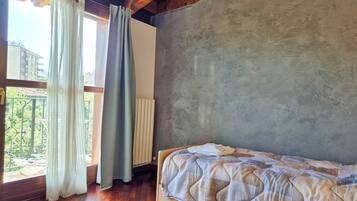 Een slaapkamer met een houten vloer, een bed met een beddegoed met dessin, een raam met gordijnen en uitzicht op groen.