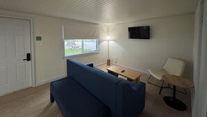 Ein Zimmer mit einem blauen Sofa, einem kleinen Holztisch, einem weißen Stuhl, einer Lampe und einem an der Wand befestigten Fernseher.