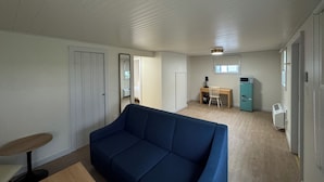 Ein Zimmer mit einem blauen Sofa, einem Schreibtisch mit Stuhl, einer kleinen Küchenzeile und einer Tür zu einem weiteren Raum.