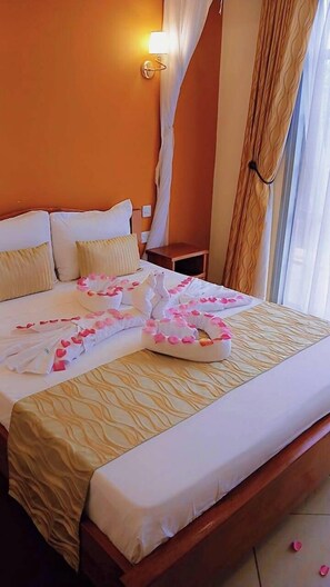 Una cama bien tendida con pétalos de rosa y un arreglo de toallas con forma de corazón, una lámpara de noche y una ventana con cortinas.