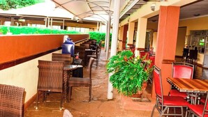 A covered outdoor seating area with wicker chairs and tables, potted plants, and a view of the building's interior. - Theta Country Resort (Juja)