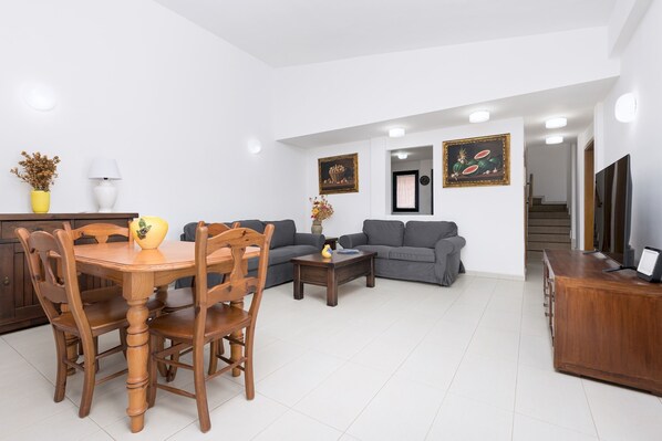 A modern living room with a dining table, chairs, a sofa, and a coffee table. There are two paintings on the wall and a TV on a wooden cabinet.