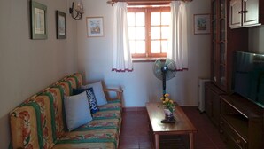 A cozy living room with a sofa, a table with a vase of flowers, a standing fan, and a TV on a cabinet.