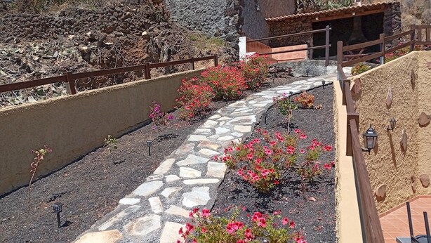A stone pathway with pink flowers, a wooden fence, and a rustic building with a tiled roof.