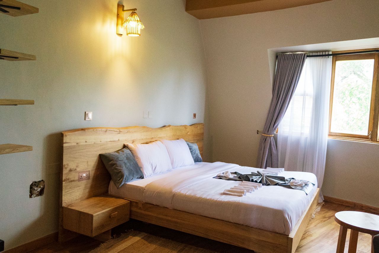 A wooden bed with a headboard, a bedside table, and a nightstand. There is a window with curtains and a wall-mounted light fixture.