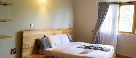 A wooden bed with a headboard, a bedside table, and a nightstand. There is a window with curtains and a wall-mounted light fixture.