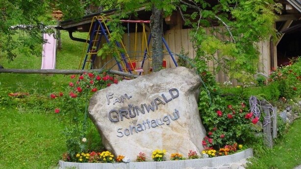A stone sign with 'Fam. Grünwald Schaffaigut' engraved, surrounded by a flower bed with various flowers.