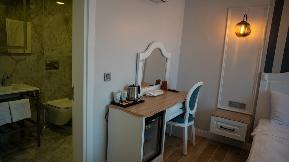 Un baño pequeño con pared de mármol, un lavamanos, un inodoro, un espejo, una silla, un mueble pequeño y una lámpara fijada a la pared.