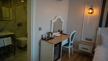 Un baño pequeño con pared de mármol, un lavamanos, un inodoro, un espejo, una silla, un mueble pequeño y una lámpara fijada a la pared.