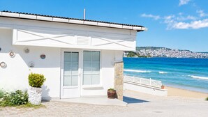 A coastal building with a white facade, a door, and windows, situated near a beach with a view of the sea and a distant cityscape. - Beyaz Ev Hotel Restaurant (Sinop)