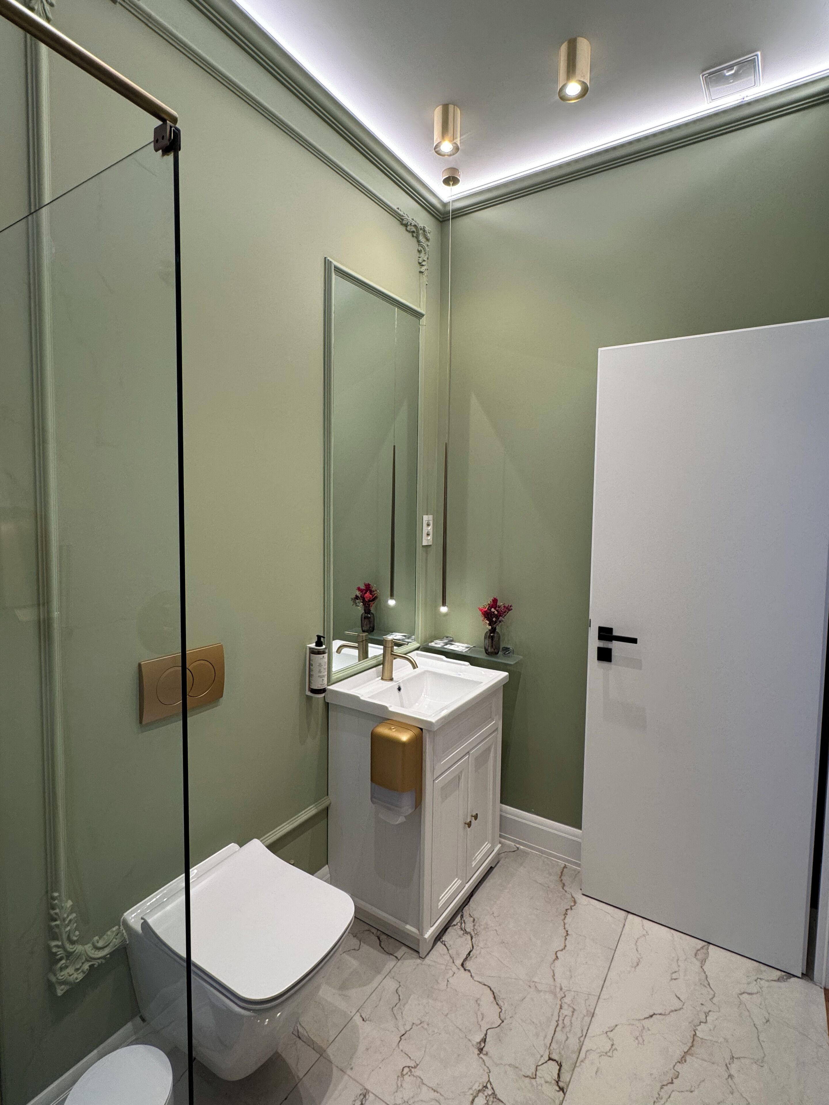 A modern bathroom with a glass shower enclosure, a white toilet, a vanity with a gold towel dispenser, and a large mirror.