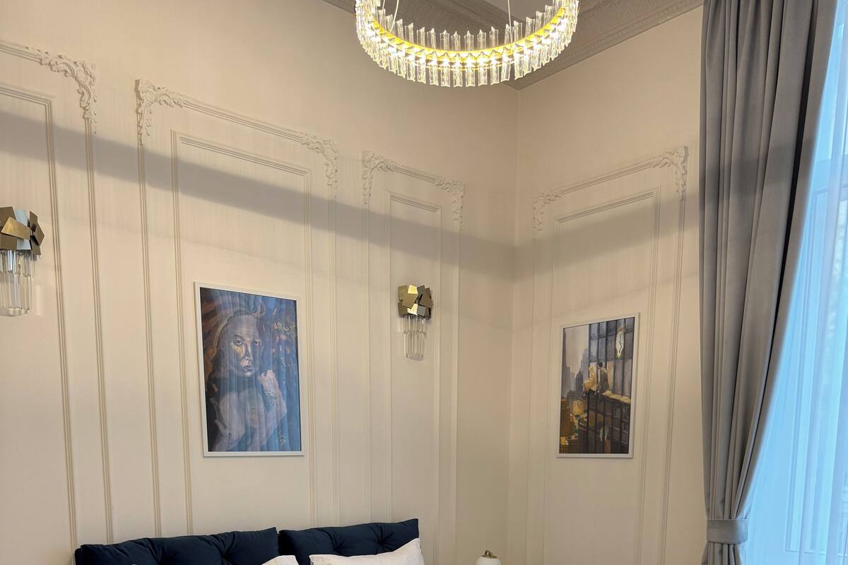 A bedroom with a large bed, white bedding, a chandelier, and two paintings on the walls.
