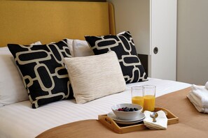 A neatly made bed with patterned pillows, a tray with breakfast including a bowl of fruit and orange juice, and a wooden headboard.
