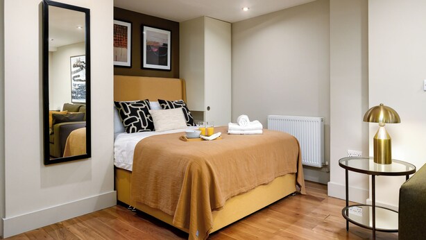 A hotel room with a bed, a round table with a lamp, a radiator, and artwork on the wall.