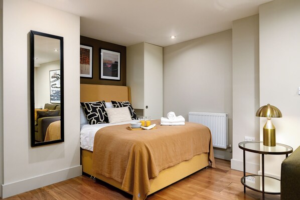 Una habitación de hotel con cama, una mesa redonda con lámpara, un radiador y una obra de arte en la pared.