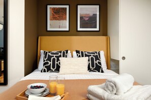 Une chambre d’hôtel avec un lit, deux œuvres d’art encadrées, un plateau de petit-déjeuner avec des fruits et du jus d’orange, et une serviette blanche roulée.