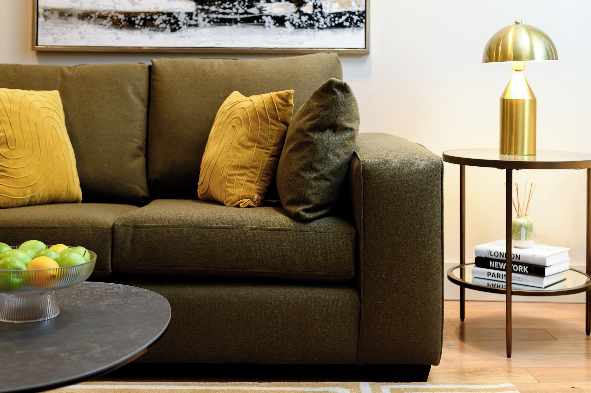 A living room with a green sofa, yellow pillows, a round coffee table, and a lamp on a side table with books.