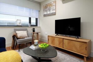 A living room with a sofa, a coffee table with books and a bowl of apples, a television, and a window with a view.