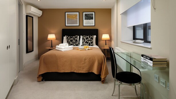 Una camera d'albergo con un letto, due lampade sui comodini, una finestra con vista, un tavolino in vetro con dei libri e una sedia.