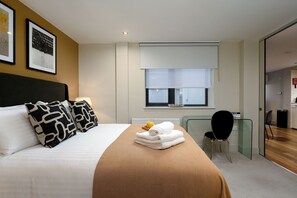 Habitación de hotel con cama, mesita de noche con vaso de jugo de naranja, toalla doblada y ventana con vista al exterior.