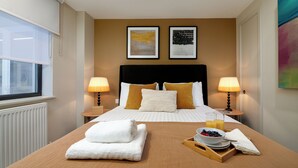 Una habitación de hotel con una cama, dos lámparas de noche, un ventanal, un radiador y una bandeja con alimentos para el desayuno.