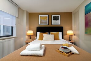 A hotel room with a bed, two bedside lamps, a window, a radiator, and a tray with breakfast items.