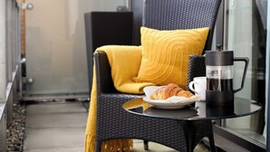 Un balcón con una silla de mimbre, una manta amarilla, un plato con un croissant, una cafetera y una taza.