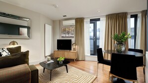 Una sala de estar moderna con televisor de pantalla plana, una mesa de centro, un sofá, un comedor con mesa y sillas, y un ventanal con cortinas.