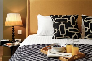 A hotel room with a bed, a nightstand with a lamp, a tray with breakfast items, and a magazine.