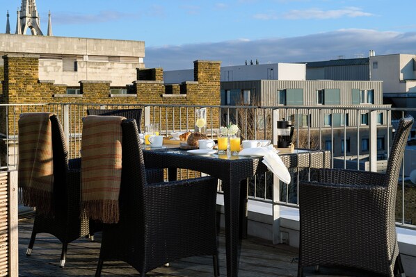 A rooftop terrace with wicker chairs, a table set for breakfast, and a view of a cityscape with modern buildings and a church spire.