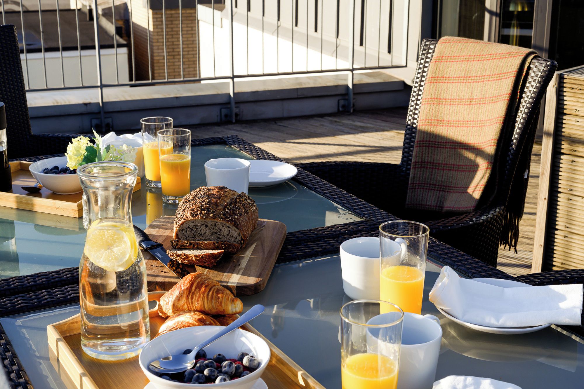 En morgenmadsopsætning med brød, croissanter og juice på et glasbord udendørs.