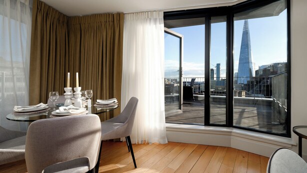 A corner room with a dining table set for two, a chair, and a large window offering a view of a city skyline.