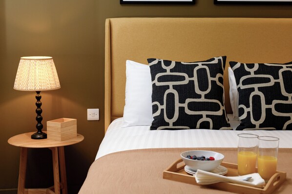 Un lit bien fait, avec un oreiller à motifs noirs et blancs, une table de chevet sur laquelle se trouve une lampe, et un plateau avec le petit-déjeuner, comprenant un bol de fruits et des verres de jus d’orange.