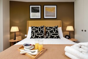 Une chambre d’hôtel avec un lit, deux tables de chevet, un plateau avec le petit-déjeuner et deux œuvres d’art mural.
