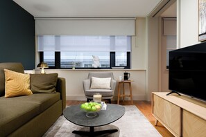 Ein Wohnzimmer mit einer Couch, einem Sessel, einem Couchtisch mit Äpfeln, einem Fernseher und einem Fenster mit Jalousien.