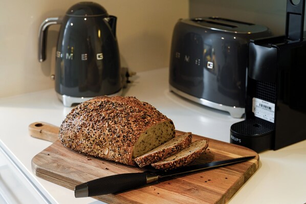 A loaf of seeded bread on a wooden cutting board with a knife, a coffee maker, and a toaster on a kitchen counter.
