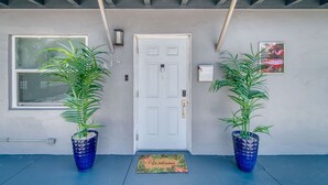 A front door with a doormat, two potted plants, a window, and a wall-mounted light fixture.