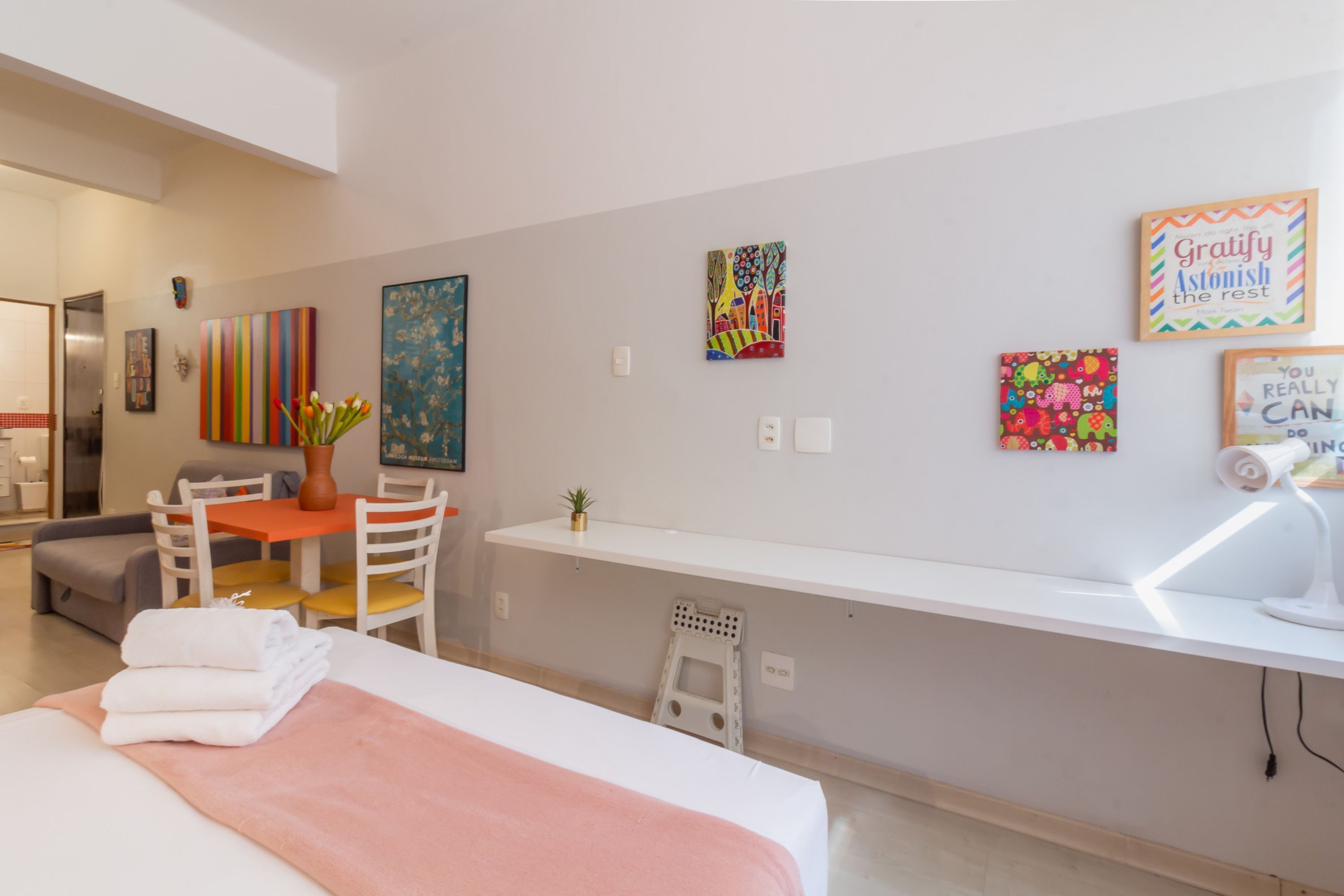 A hotel room with a bed, a desk, a chair, a small table, and a potted plant. There are several artworks on the wall.