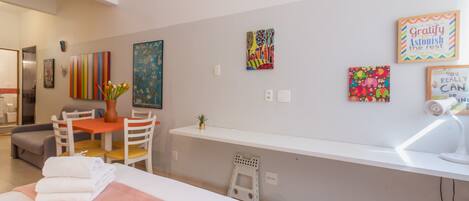 A hotel room with a bed, a desk, a chair, a small table, and a potted plant. There are several artworks on the wall.