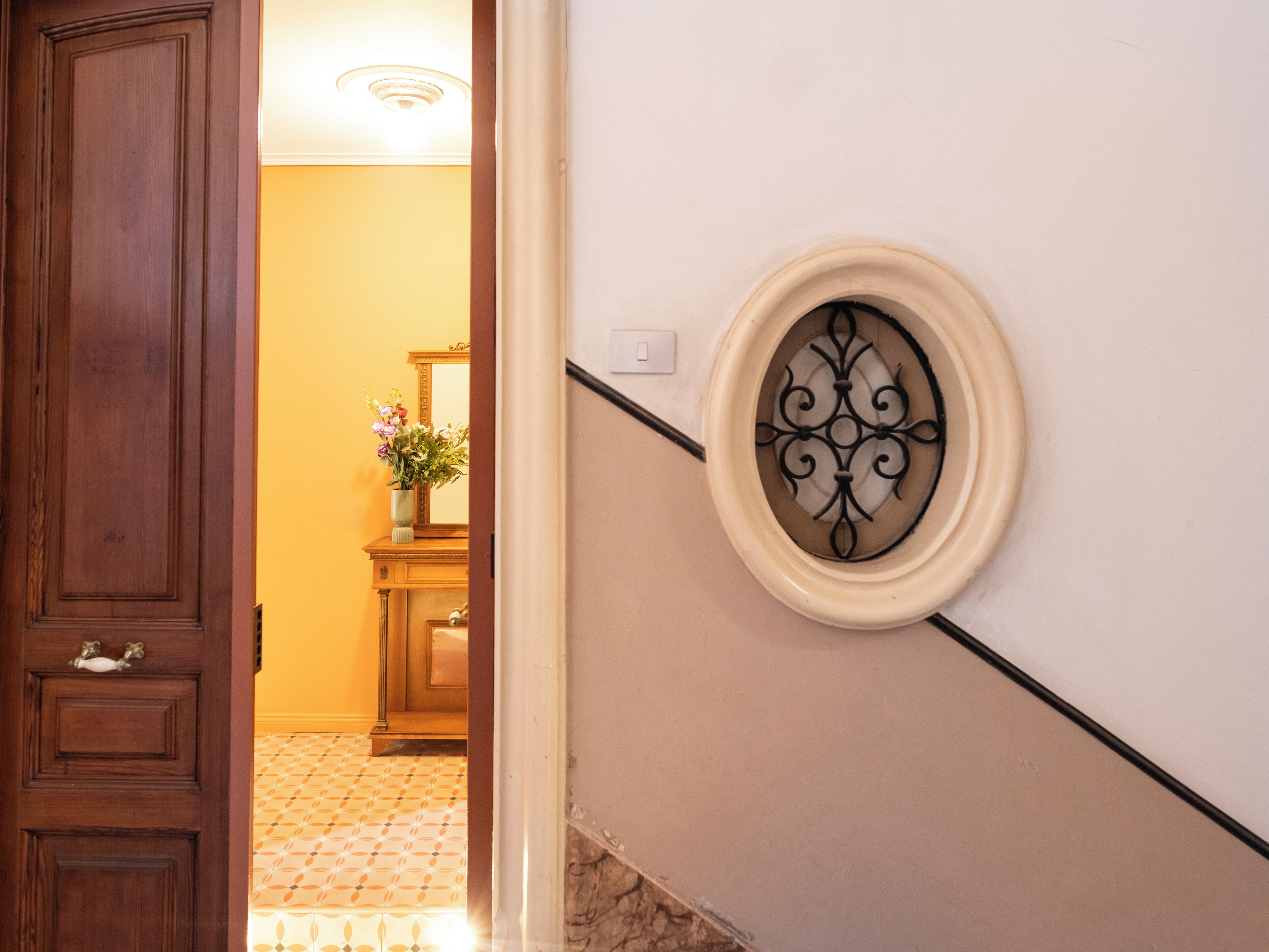 Un pasillo con puerta de madera, un espejo, un jarrón con flores y un plato decorativo en la pared.