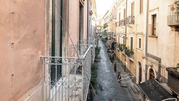 Una calle estrecha flanqueada por edificios, con balcones que lucen barandillas y plantas en macetas.
