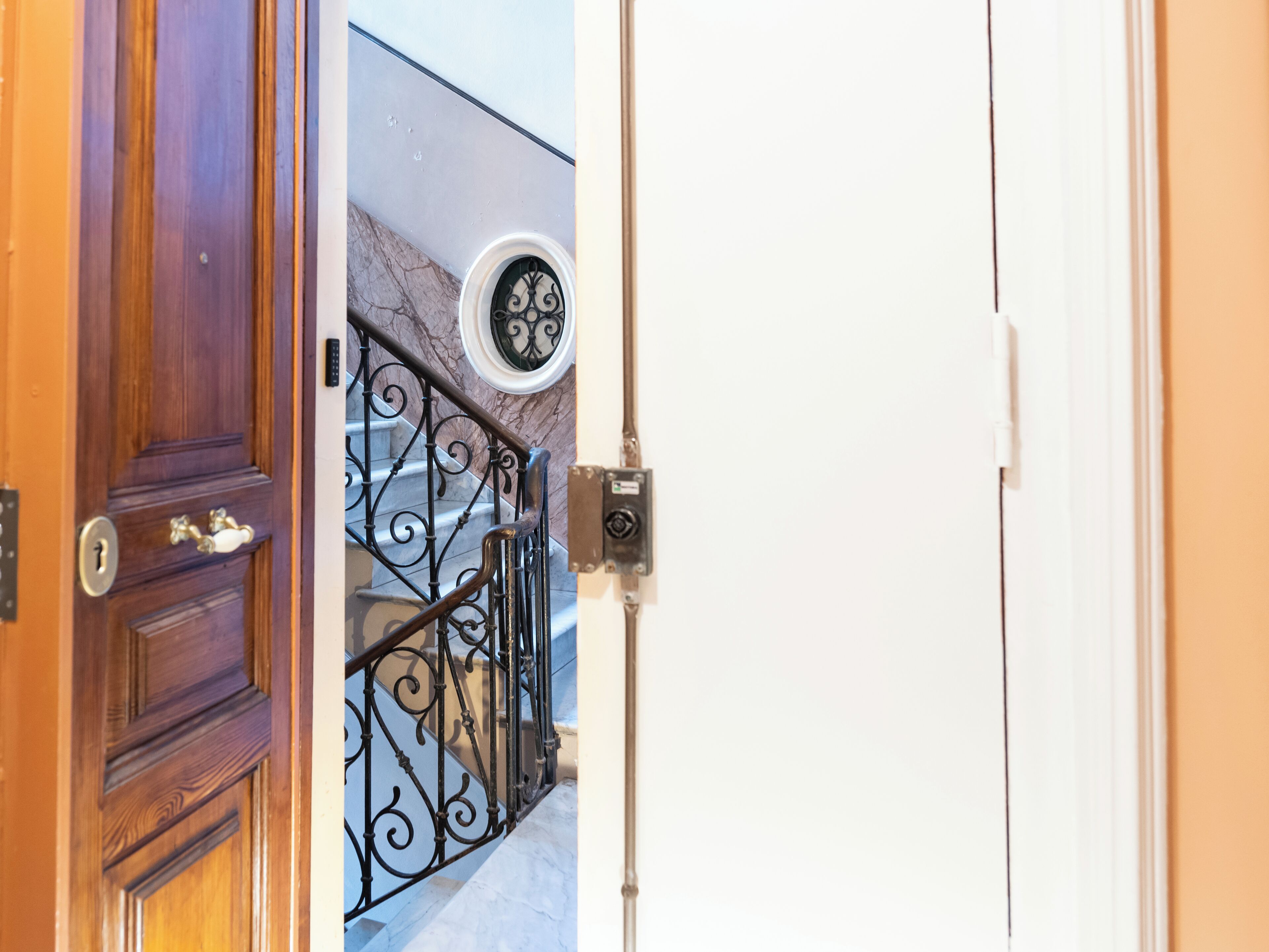 Una puerta de madera con un ojo de cerradura y un pomo de latón, que da a una escalera con barandilla de hierro forjado negro y un ventanal circular.