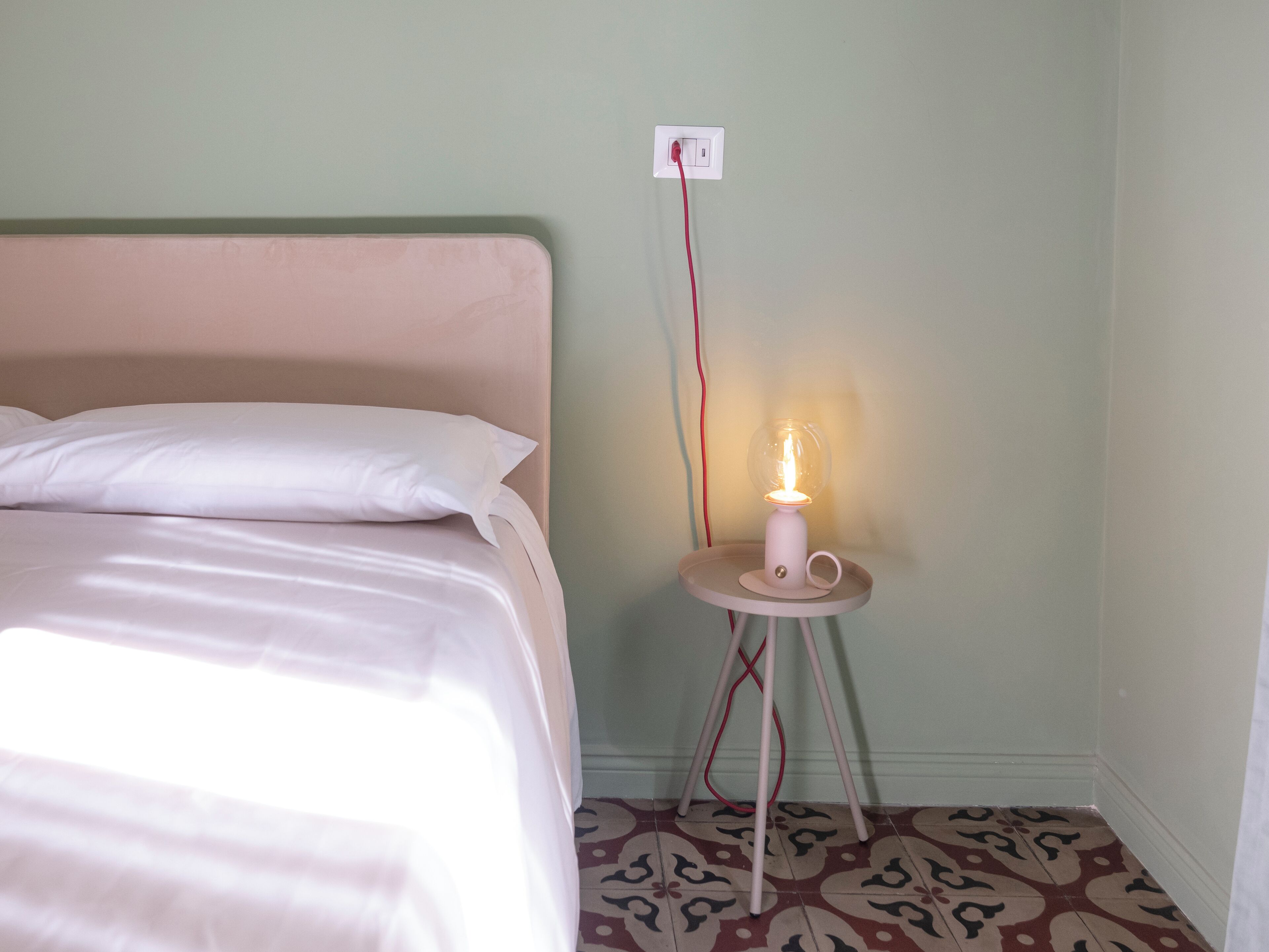 Una cama con ropa de cama blanca, una mesita de noche con lámpara y un enchufe en la pared.