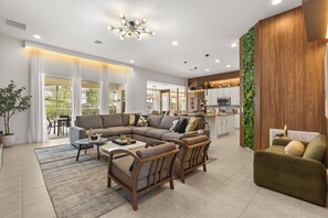 Ein modernes Wohnzimmer mit Ecksofa, zwei Sesseln, einem Couchtisch und einem Essbereich mit Tisch und Stühlen. Es gibt eine Küchenzeile mit Holzschränken und einer vertikalen grünen Wand.