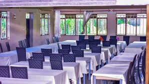 A dining area with multiple tables and chairs, large windows with bars, and a view of greenery outside.