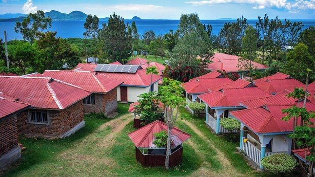 A coastal resort with red-roofed cottages, a grassy courtyard, and a view of the sea and distant mountains.