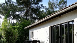 A wooden deck with a table and chairs, a white building with large windows, and surrounding trees. - Villa on the tip of the CAP FERRET peninsula (CAP FERRET)