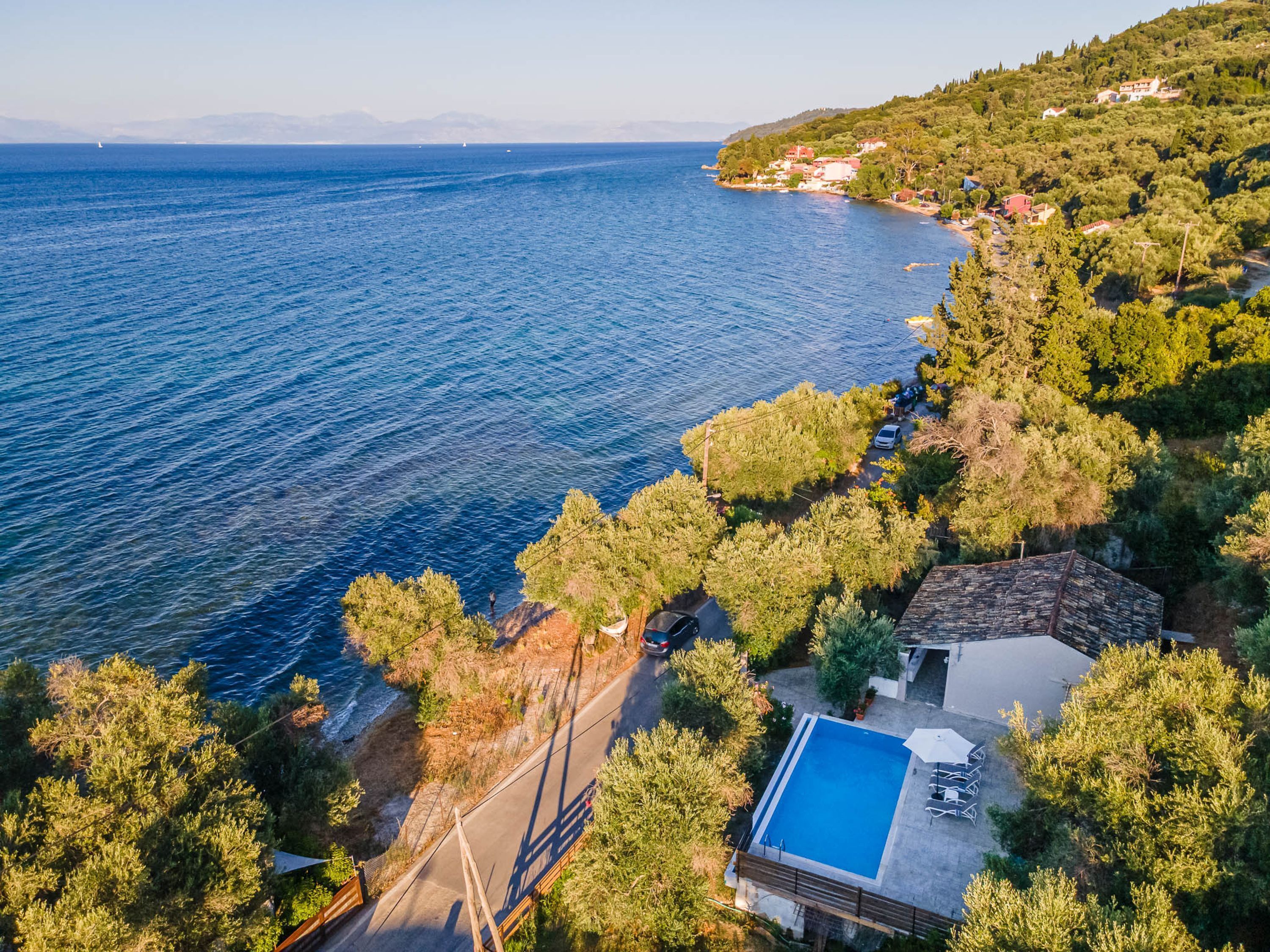 An aerial view of a coastal property with a swimming pool, a road, and surrounding greenery.