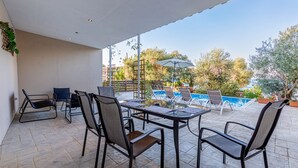 An outdoor patio with a table and chairs, a pool area with lounge chairs, and a view of trees and a building in the background. - Deja Blue Beach Pool House by Estia (Corfu)