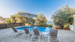 A swimming pool with lounge chairs and an umbrella, surrounded by trees and a view of the sea. - Deja Blue Beach Pool House by Estia (Corfu)